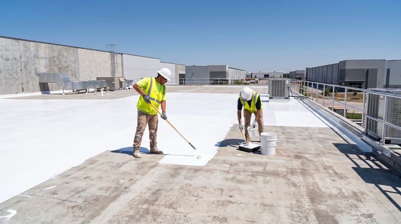 Two commercial roofing workers applying a white elastomeric reflective roof coating with paint rollers on a flat industrial building roof to improve energy efficiency and seal the surface on a clear - Oak Grove Roofing